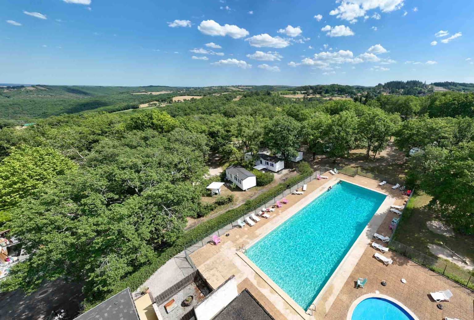 Vue aérienne du camping Le Picouty dans le Lot : grande piscine extérieure, mobil-homes sous les chênes et collines boisées à perte de vue
