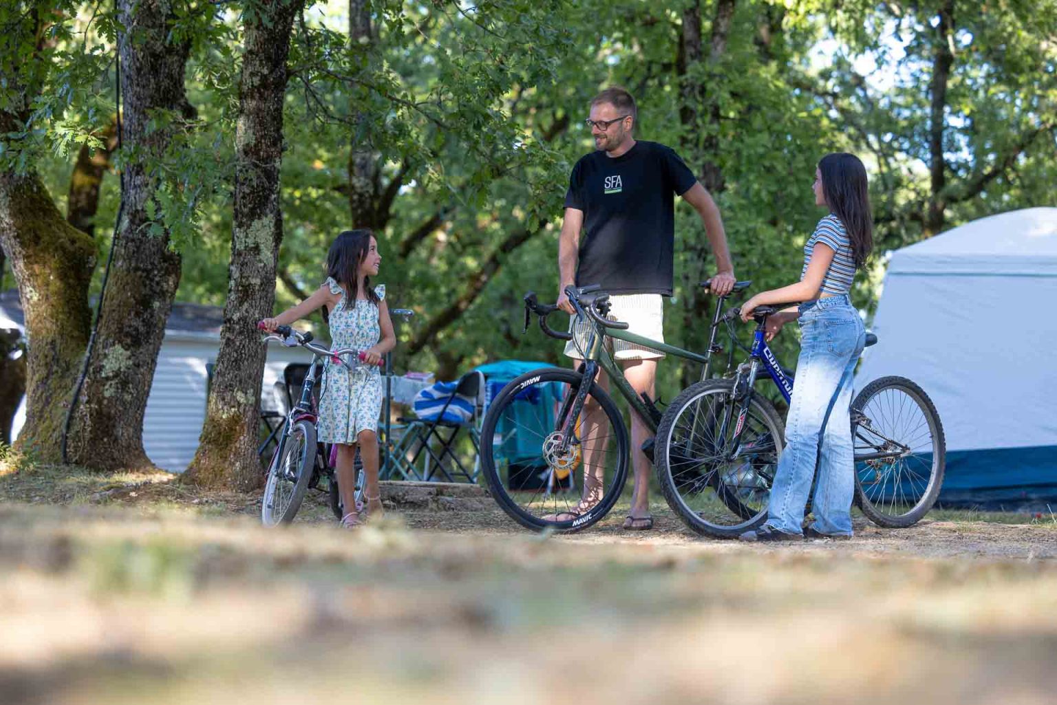 Famille avec ses vélos sur un emplacement de camping ombragé sous les chênes, tente plantée en arrière-plan