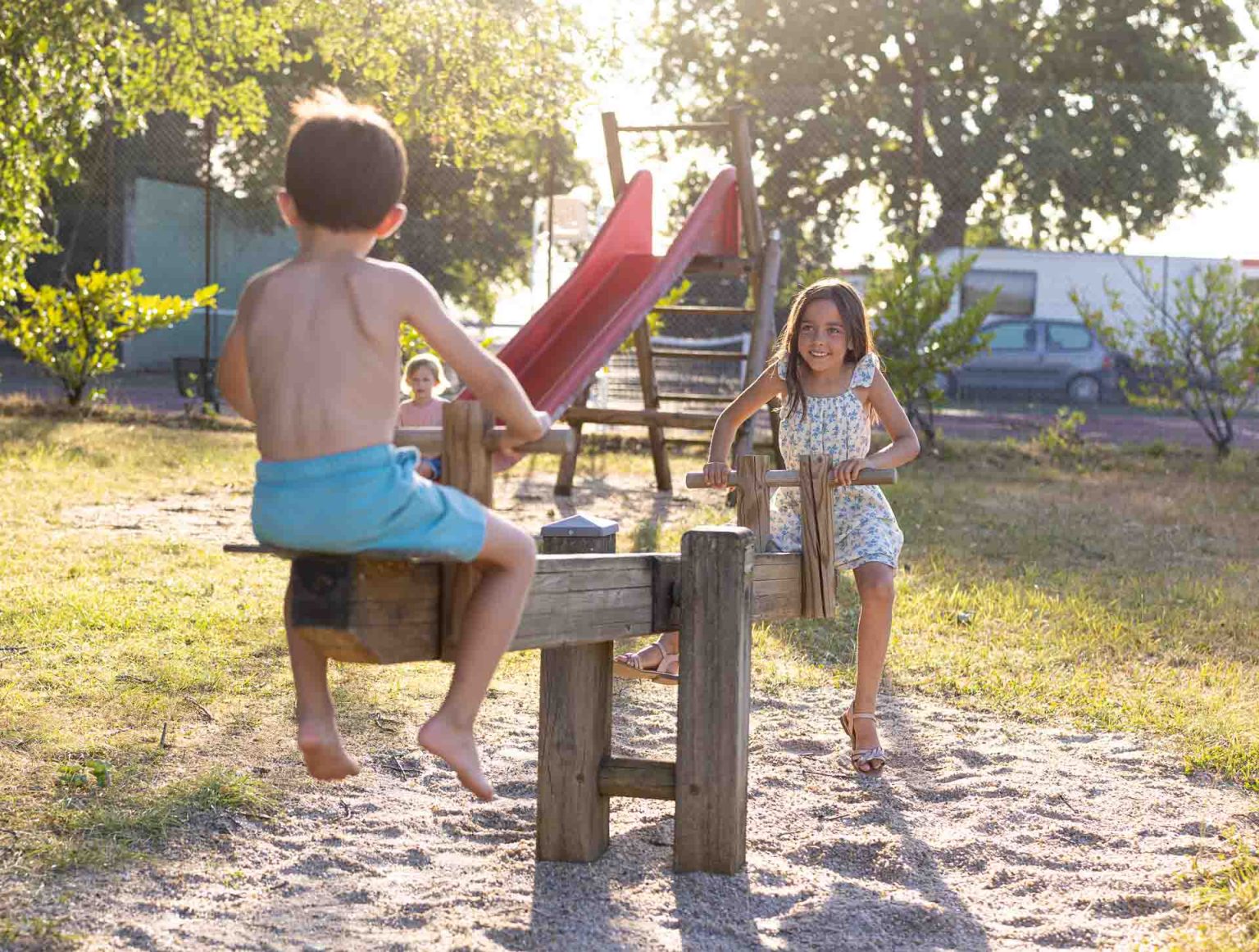 Deux enfants jouent à la bascule en bois sur l’aire de jeux du camping, toboggan et soleil couchant en arrière-plan