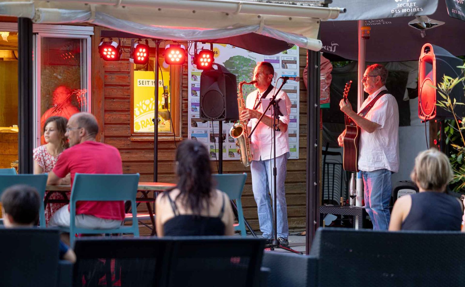 Concert en terrasse au bar du camping : saxophoniste et guitariste en soirée devant les vacanciers attablés